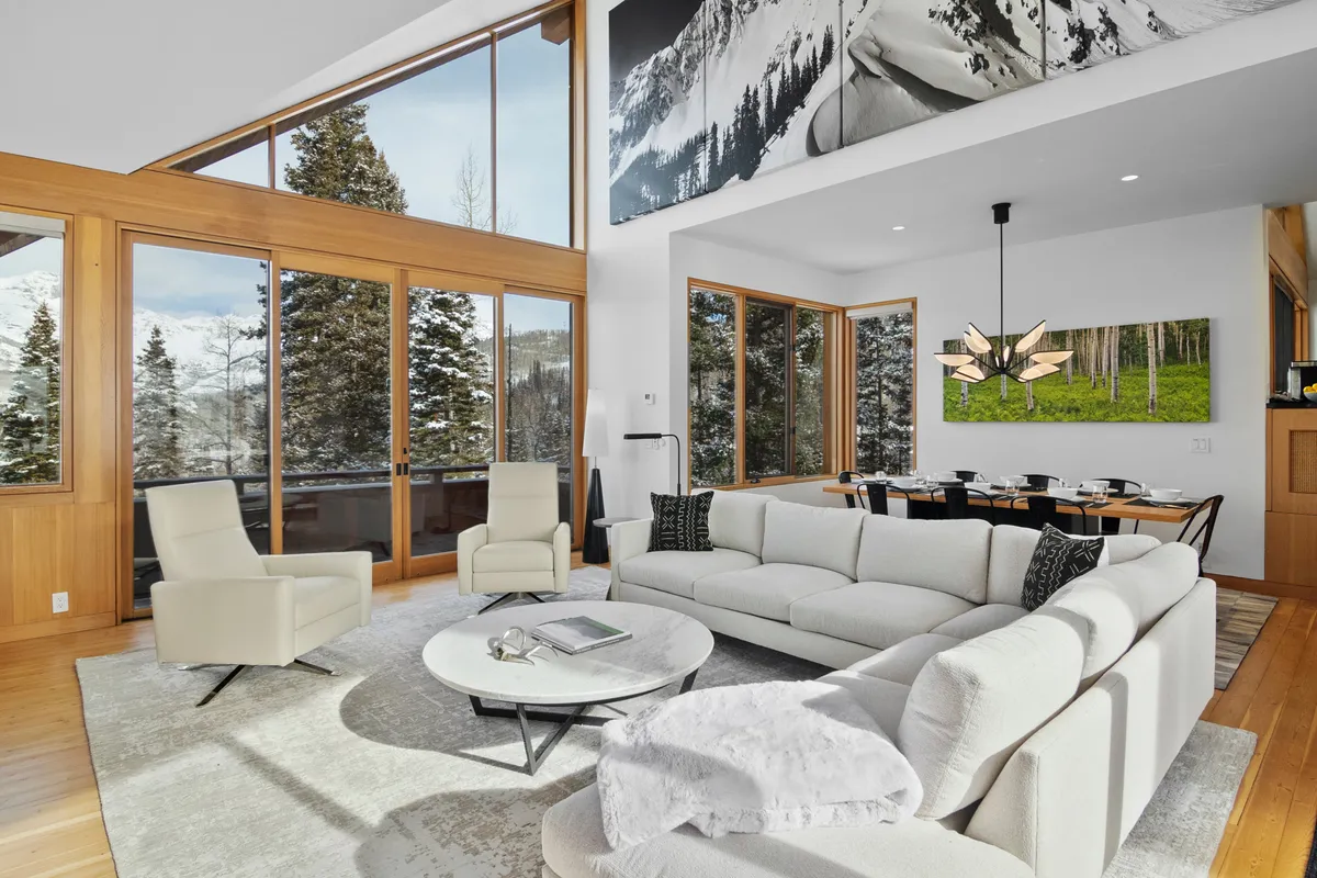 A spacious living area with a modern sectional sofa faces soaring floor-to-ceiling windows that reveal dramatic mountain views. The room is filled with cozy yet contemporary furnishings, eye-catching artwork, and a unique light fixture. The open layout suggests an easy flow to adjacent dining or kitchen spaces, while the panoramic glass amplifies the connection to the surrounding landscape.