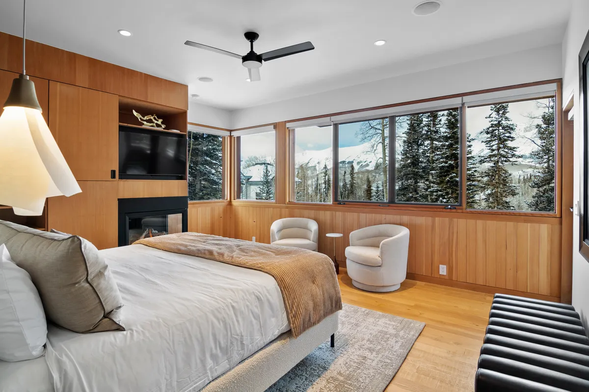 A spacious bedroom with soft linens on the bed, modern armchairs, a welcoming fireplace, and expansive windows framing breathtaking mountain views. The decor is tranquil and sophisticated, emphasizing peaceful seclusion.