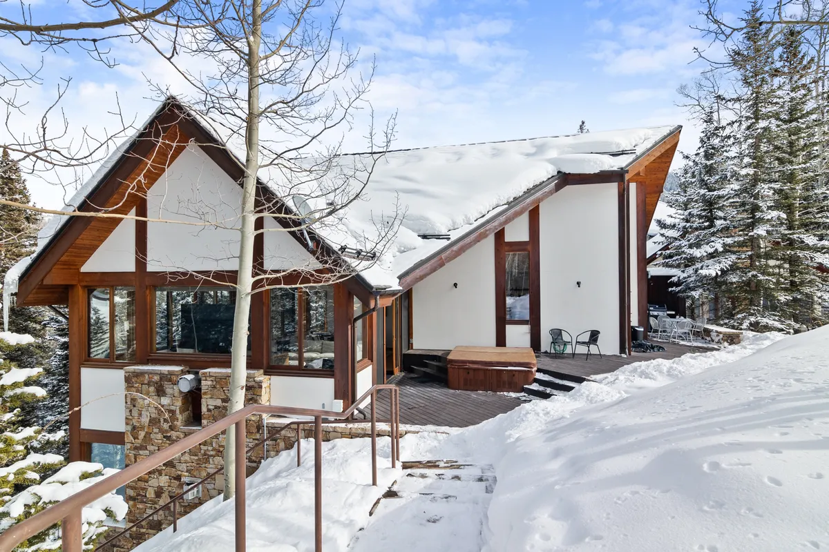 A modern architectural exterior shot featuring the chalet’s snow-covered roof, a wooden deck, and mature trees. The inviting deck suggests spaces for outdoor gatherings, while the surrounding snow hints at both winter adventure and cozy togetherness.