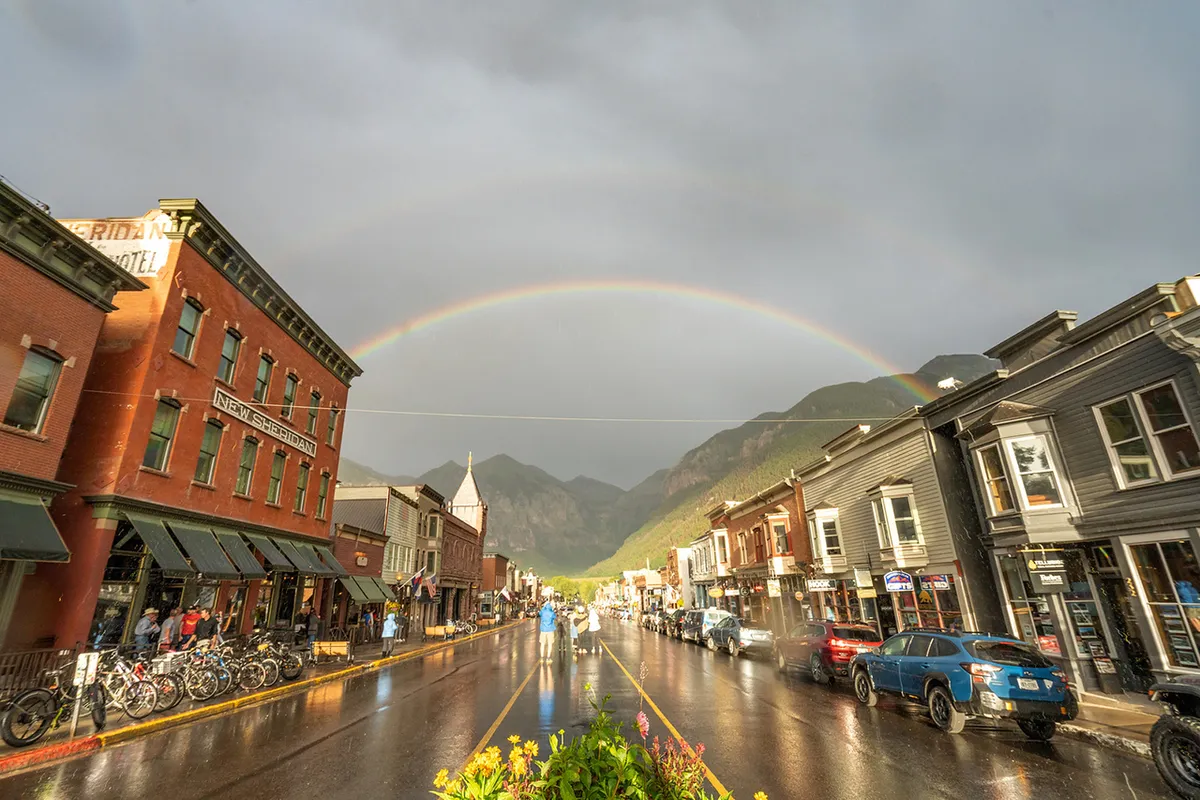 A vibrant photo of Telluride’s historic core on a summer day, featuring colorful buildings, bicycles, a rainbow, and majestic mountains in the background. The image captures people exploring, shopping, and dining outdoors.