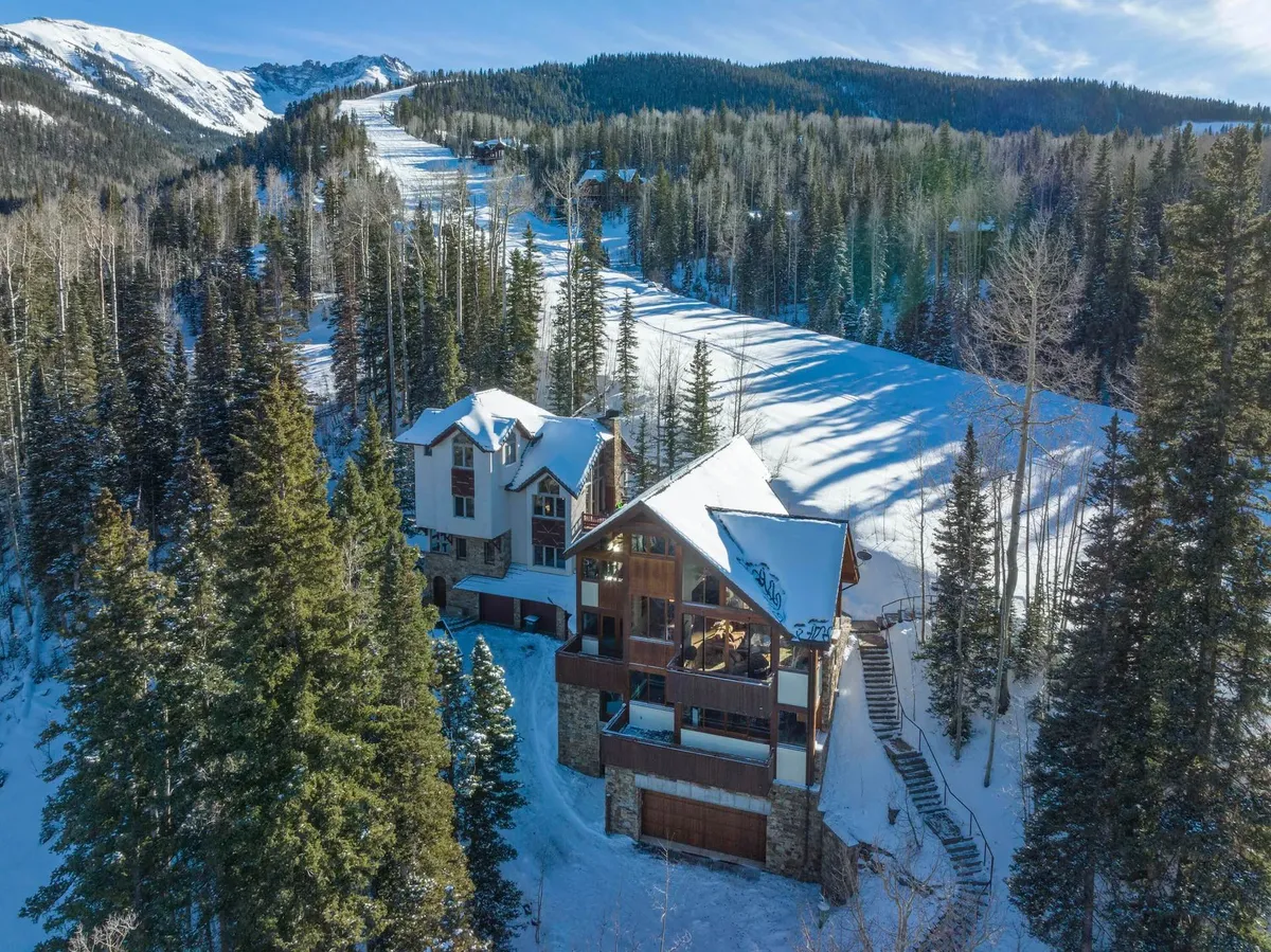 A spacious mountain home nestled among snow-covered trees, with the ski slope visible in the background. The setting conveys a serene winter landscape and hints at the home's direct access to the slopes.