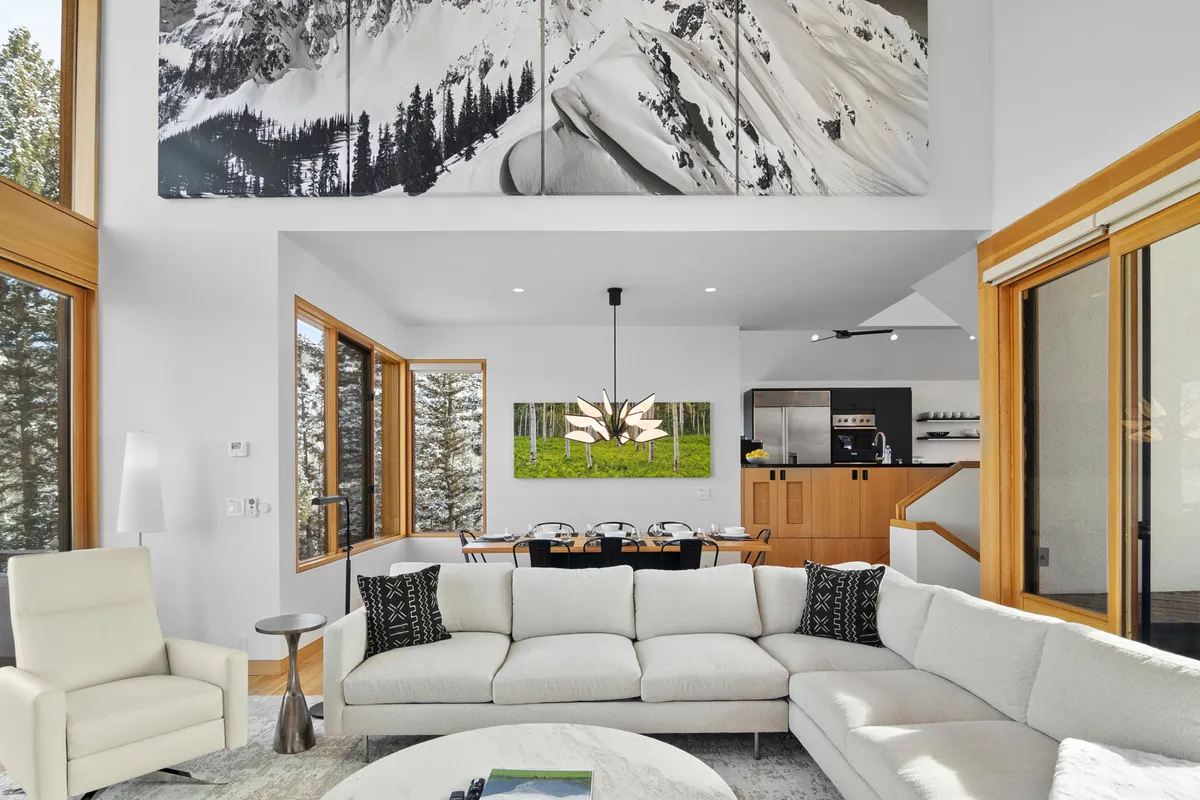 A view of the open-plan living and dining area, featuring a large sectional sofa, modern dining table with chairs, a striking light fixture, and bold mountain artwork. Ample daylight pours in from the large windows, fostering a bright and welcoming atmosphere ideal for gatherings.