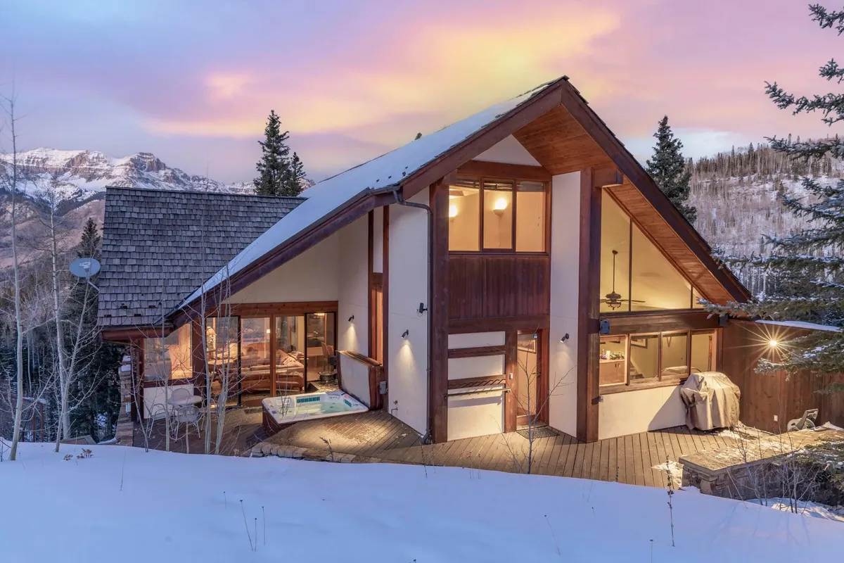A modern chalet exterior with an inviting hot tub on the deck, surrounded by mountains and forest. The home’s architecture draws the eye to outdoor amenities and panoramic views, suggesting a space for rejuvenation after summer adventures.
