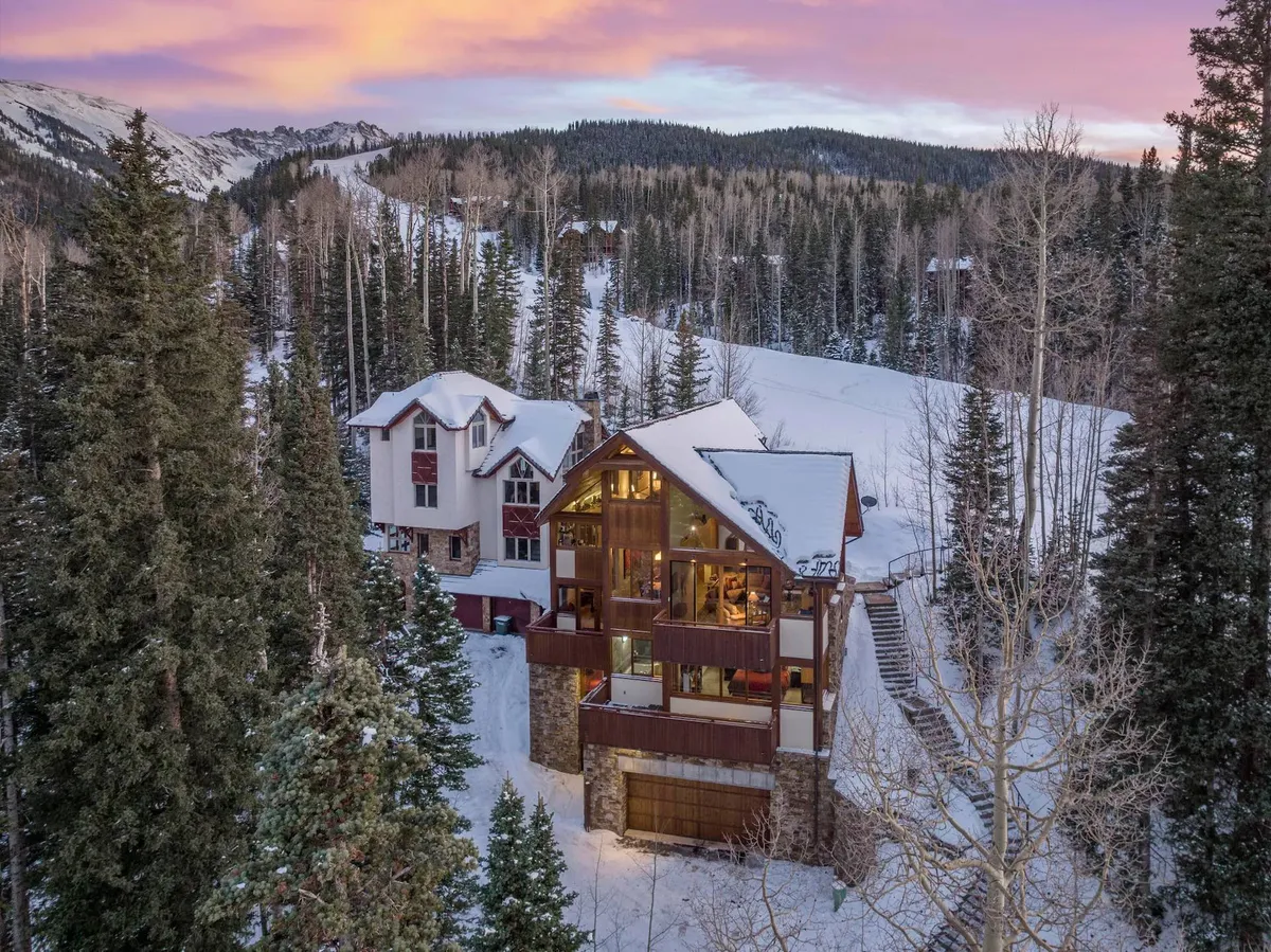 A welcoming view of the main house set against a snowy landscape, with the roof blanketed in snow and tall trees surrounding the property. The scene conveys warmth and a sense of arrival, inviting families and groups to gather in a picturesque winter setting.