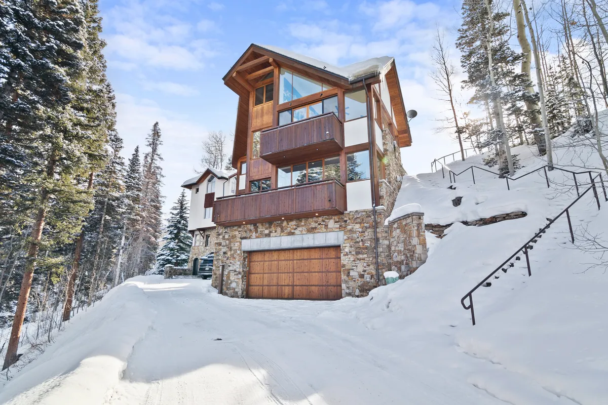 A striking exterior view of the home showcases its modern architectural design, with a stone facade, expansive snow-covered driveway, and tall pine trees framing the property. The scene captures a sense of arrival, highlighting the relationship between the home's design and its breathtaking natural surroundings.