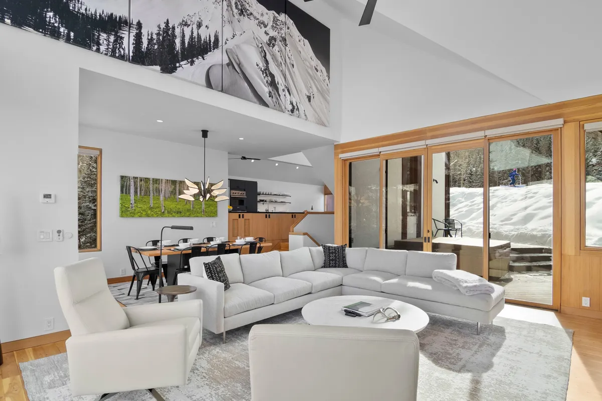 The image showcases a spacious, contemporary open-plan living and dining area with a large, plush sectional sofa facing a wall adorned with dramatic mountain artwork. Sunlight pours in through generous windows, illuminating the cozy yet modern space, while the adjacent dining area and wooden cabinetry hint at a welcoming environment for gatherings. The overall atmosphere is both inviting and luxurious, perfect for relaxation or entertaining after a day on the slopes or trails.