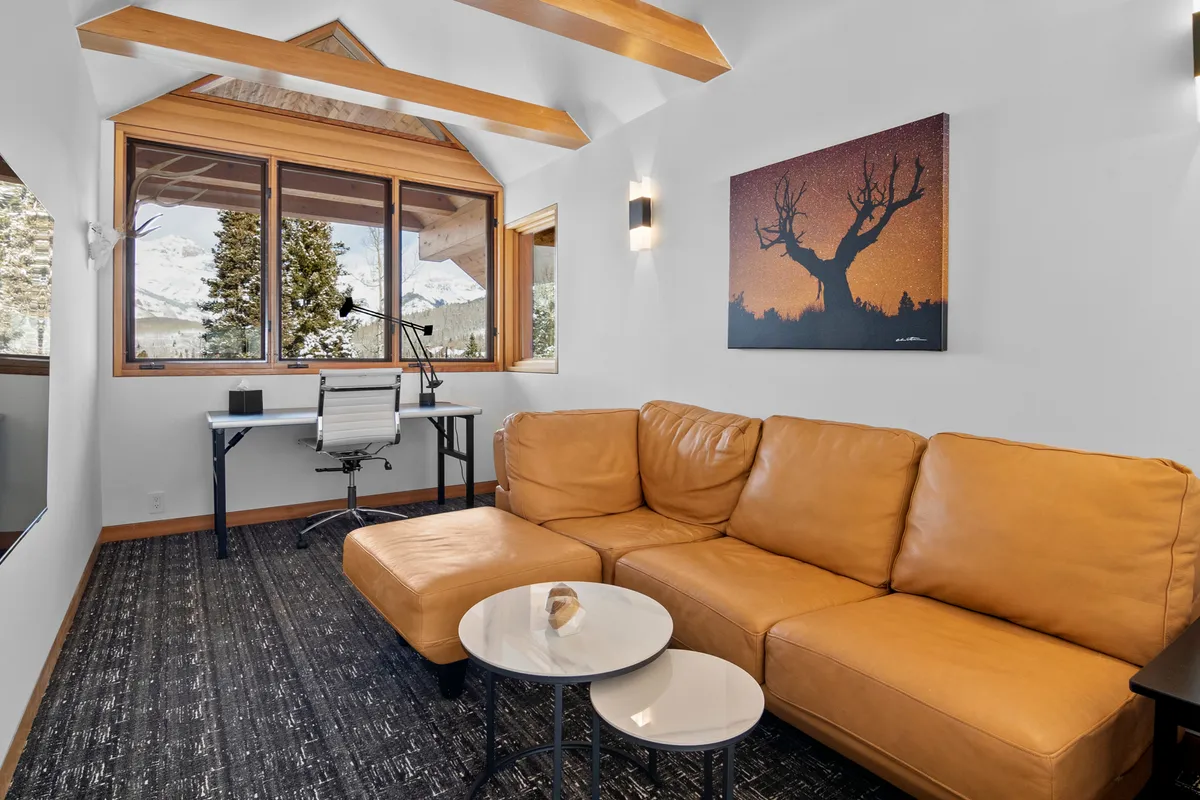 A comfortable loft space featuring a leather sectional sofa, accent tables, and a desk set by a large window. The scene is both cozy and functional, capturing the flexibility for children's movie nights or remote work while still enjoying the mountain views.