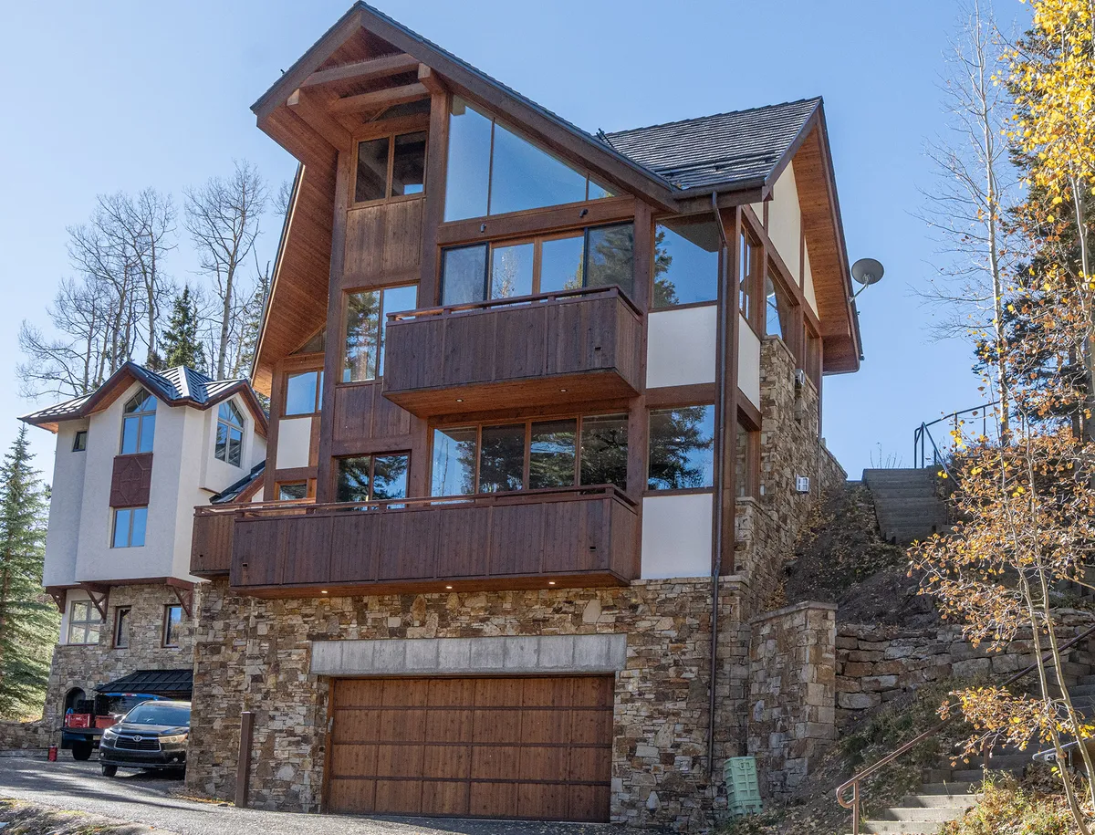 This image shows the exterior of the home with large glass windows, a wooden balcony, and a stone facade. The scene includes steep stairs leading up to the entrance, surrounded by trees, emphasizing the connection to nature and outdoor relaxation spaces.